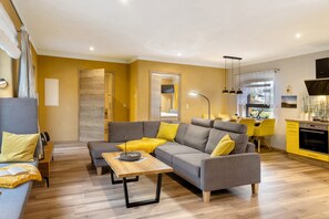 A modern living room with a grey sofa, yellow cushions, a wooden coffee table, and a kitchen area with yellow cabinets. - Holiday Home 'Mond am See' with Lake View, Private Terrace and Wi-Fi (Stechow-Ferchesar)