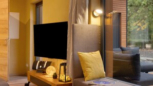 Una sala de estar moderna con un televisor de pantalla plana, una mesita de madera, un sofá con cojines y un ventanal con vistas a árboles.
