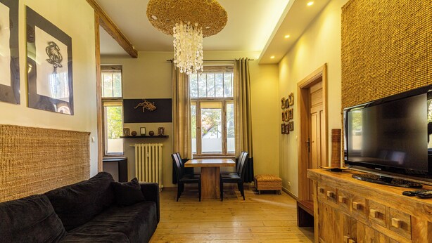 A modern living room with a dark-colored sofa, a wooden dining table with chairs, a television on a wooden cabinet, and a large window with curtains. - Africana by Grand Apartments (Sopot)