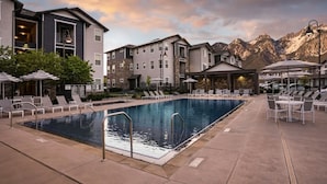 A pool area with lounge chairs and umbrellas, surrounded by buildings with stone facades and a mountainous backdrop.