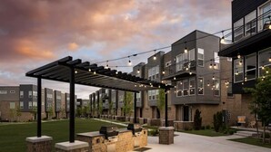 A covered outdoor patio with grilling stations, a pergola, and string lights against a backdrop of modern apartment buildings.