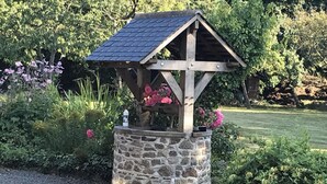 A stone structure with a wooden roof and a small platform, surrounded by greenery and flowers. - Countryside Home Sleeps 6 - Garden, Parking, Wifi (Landivy)