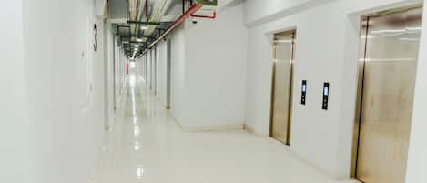 Un largo pasillo bien iluminado con pisos y paredes blancos y brillantes, dos ascensores, y tuberías y conductos del techo a la vista.