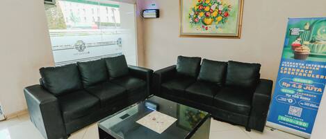 Un área de recepción con dos sofás de cuero negro, una mesa de centro de vidrio, un cuadro enmarco con motivos florales, una unidad de aire acondicionado y un cartel en la pared.