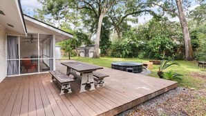 Une terrasse en bois avec une table de pique-nique et des bancs, entourée d’un jardin luxuriant et d’arbres.