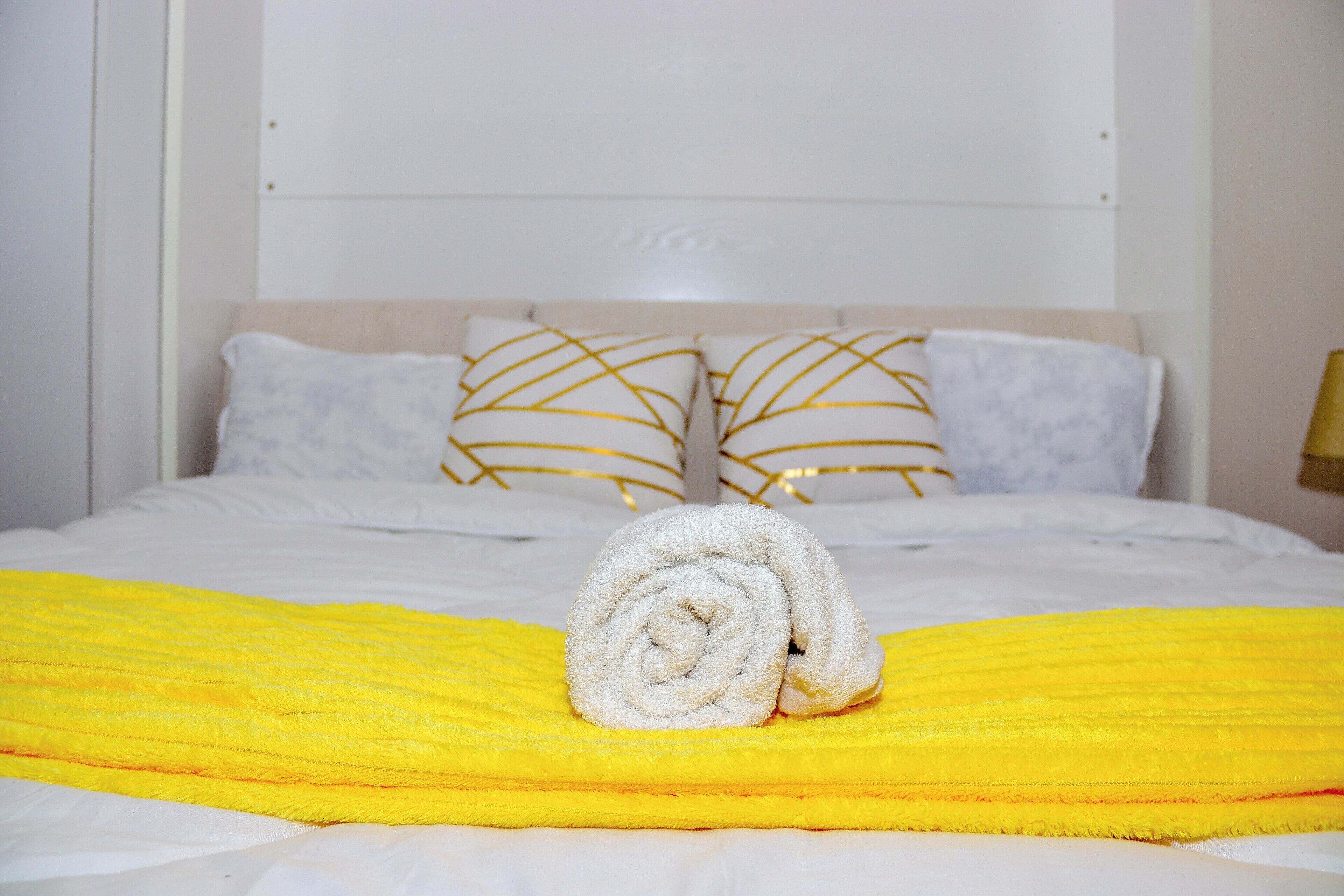 A neatly made bed with a rolled white towel and a yellow blanket, two white pillows with gold stripes, and a white headboard.