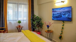 Habitación de hotel con una cama, cortinas, una planta en maceta, una mesita con un florero y un televisor de pared mostrando una imagen panorámica.