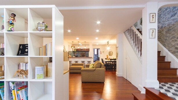 A spacious living area with a wooden floor, a sofa, a bookshelf, and a staircase.