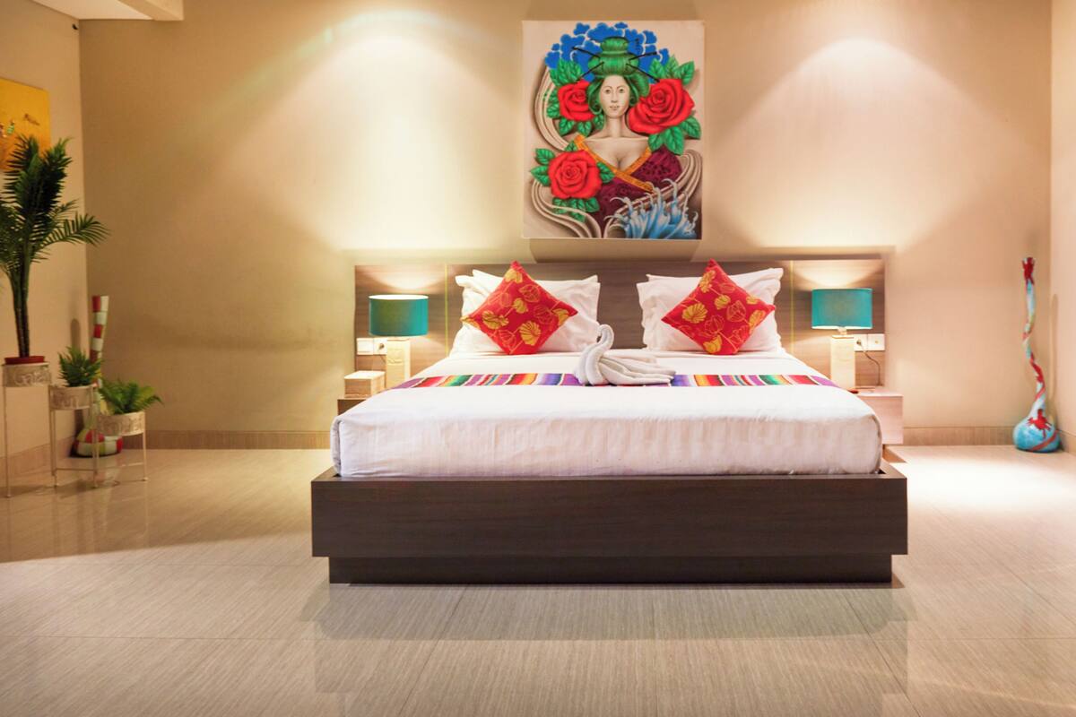 A modern hotel room with a large bed, two bedside tables, a painting above the bed, and a vase on the right side.