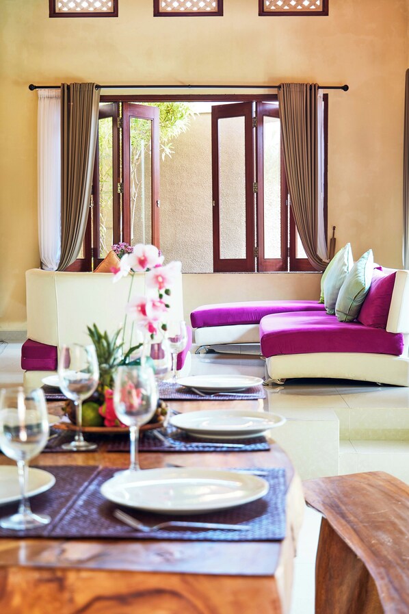 A dining area with a table set for two, a sofa with cushions, and a view of the outdoors through a glass door. - Dreamscape Bali Villa (Canggu)