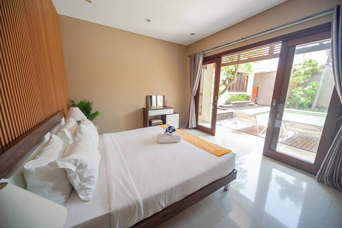 A hotel room with a large bed, a small table, a chair, and a view of the outdoor area.