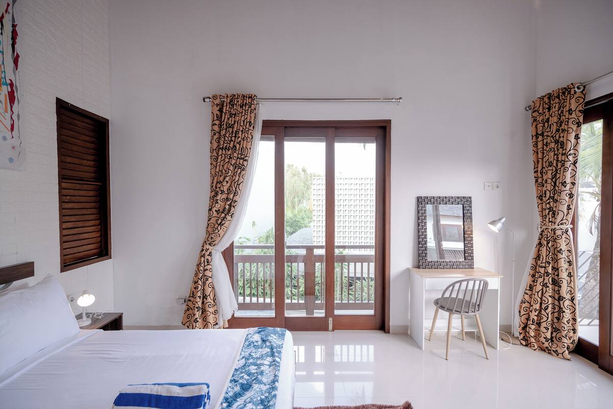 A bedroom with a bed, a desk, and a chair. There is a window with curtains and a view of the outside.