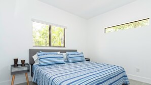 Ein modernes Schlafzimmer mit einem großen Bett, einem Nachttisch, einem Fenster mit Jalousien und einem kleinen Fenster über dem Bett.