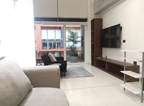Uma sala de estar moderna com sofá, televisão e varanda com vista para um edifício e palmeiras.