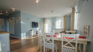 A dining area with a white table and chairs, a television on the wall, and a kitchen in the background. - Nautica by Grand Apartments (Sopot)