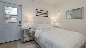 A bedroom with a bed, two bedside lamps, a framed picture on the wall, and a door with a view of palm trees.
