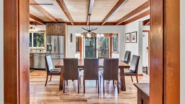 Un coin repas avec une table et des chaises en bois, un ventilateur de plafond et une vue sur la cuisine et l’extérieur.