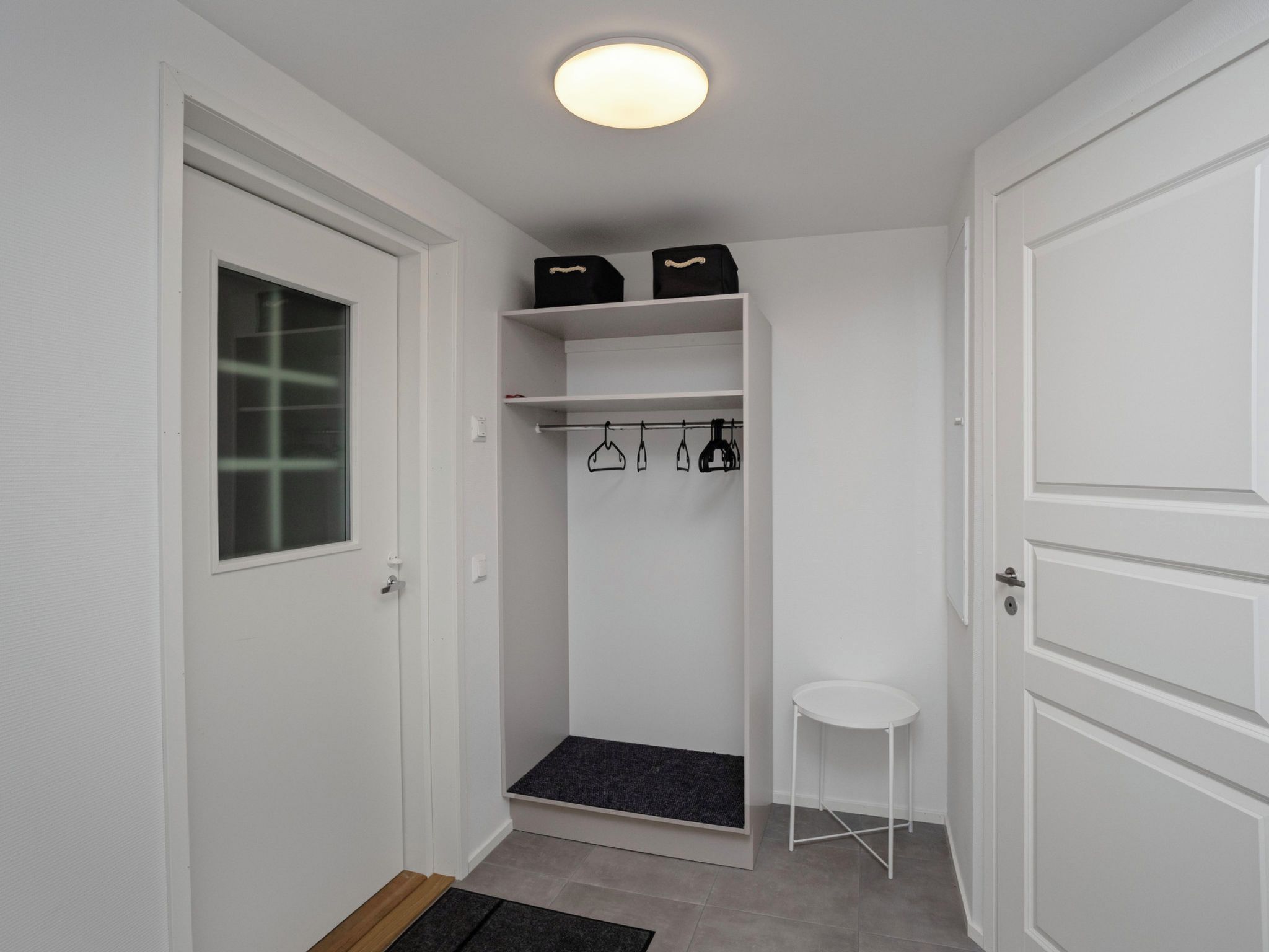 A small, white-walled room with a door, a window, a shelf with storage boxes, a hanging rack with bags, and a small white stool.