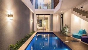 A modern indoor pool area with a glass door leading to a bedroom, a staircase, and a seating area with cushions. - Berawa Bali Beach Villa (Canggu)