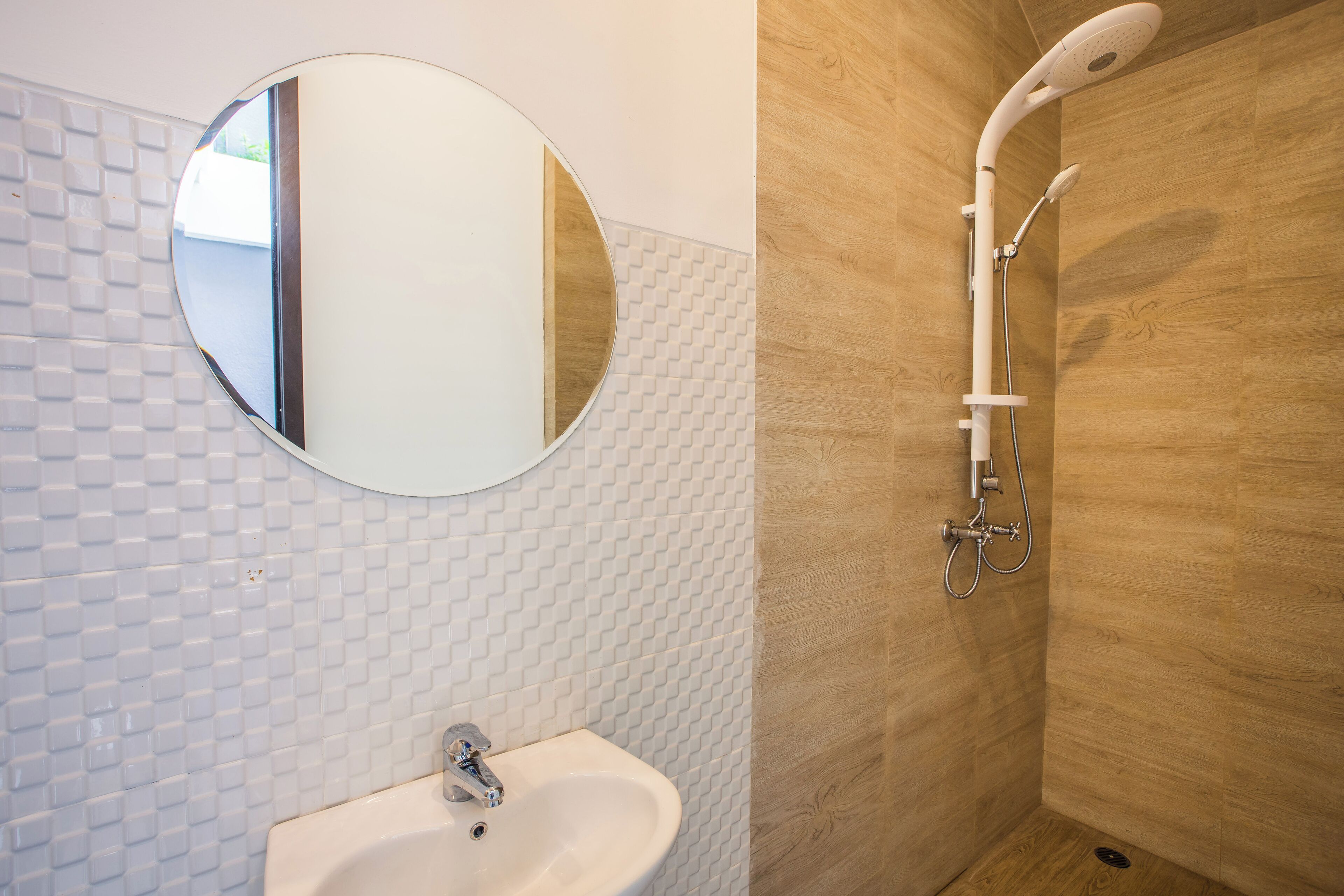 A bathroom with a white tiled wall, a round mirror, a sink, and a shower with a wooden wall.