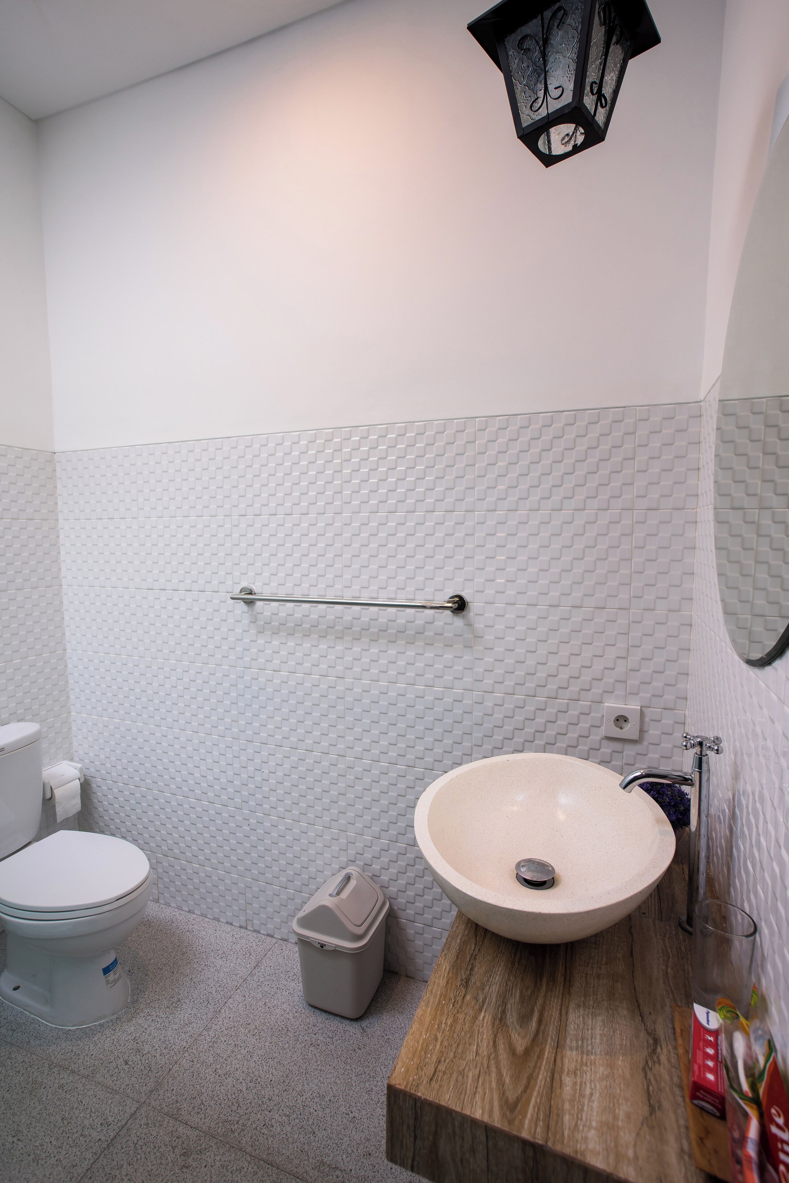 A bathroom with a white tiled wall, a wooden vanity, a round sink, a toilet, a trash bin, a towel rack, and a wall-mounted light fixture.