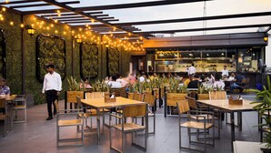 An outdoor dining area with wooden tables and chairs, string lights, and a green wall with plants.