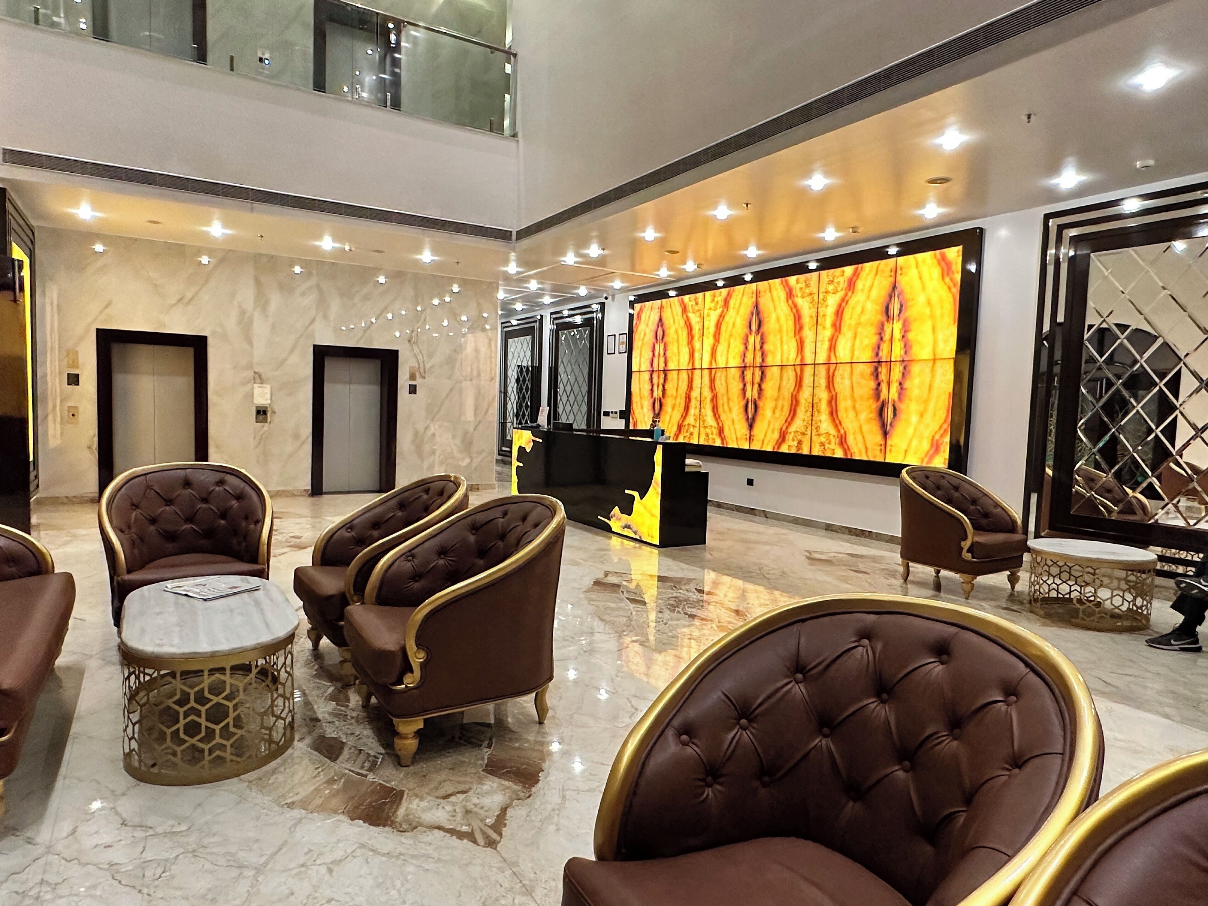 A lobby with a high ceiling, marble flooring, and a large wall-mounted screen displaying a fiery pattern. There are several upholstered chairs with gold trim and a small round table.