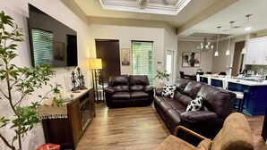 Ein modernes Wohnzimmer mit einer Couch, Sesseln und einem Couchtisch. Es gibt einen Essbereich mit Tisch und Stühlen sowie eine Küche mit einer blauen Kochinsel.