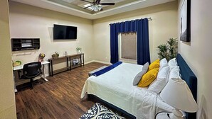 Ein Schlafzimmer mit Bett, Schreibtisch, Stuhl, Deckenventilator, Fernseher und einem Fenster mit Vorhängen.