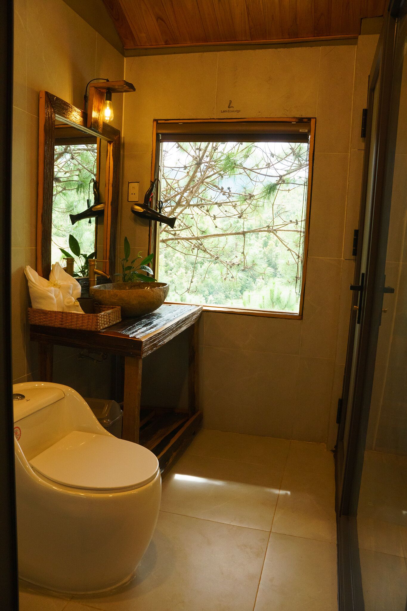 A bathroom with a toilet, a sink with a wooden countertop, a large window with a view of trees, and a mirror with a light fixture.