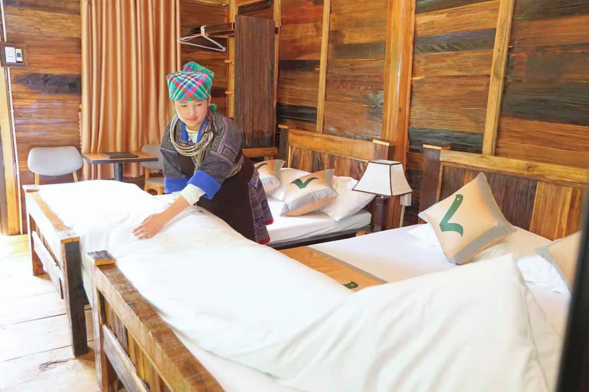 A wooden room with two beds, each with white linens and pillows, and a person arranging the bed linens.