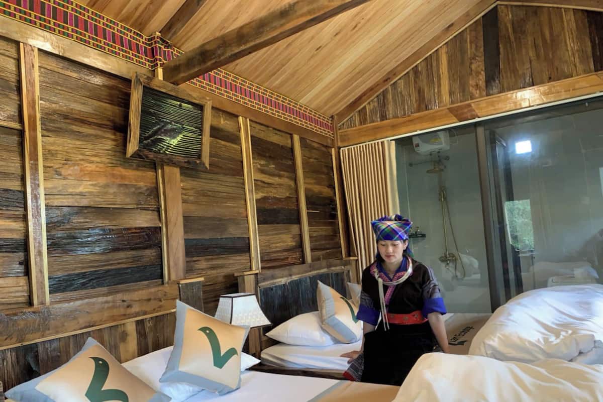 A wooden room with a bed, pillows, a fan, and a person standing by the bed.