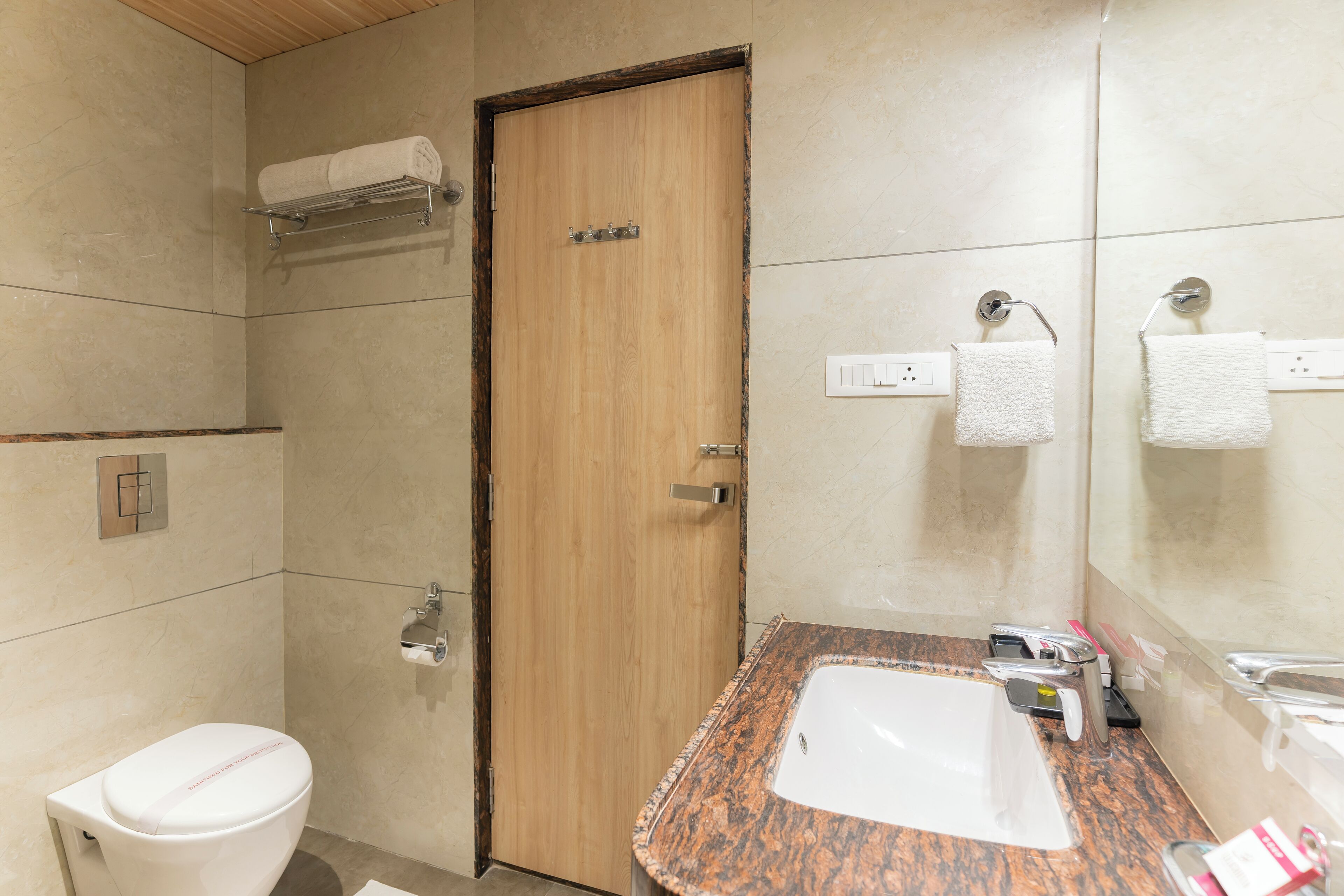 A bathroom with a wooden door, a white toilet, a sink with a granite countertop, and a mirror.