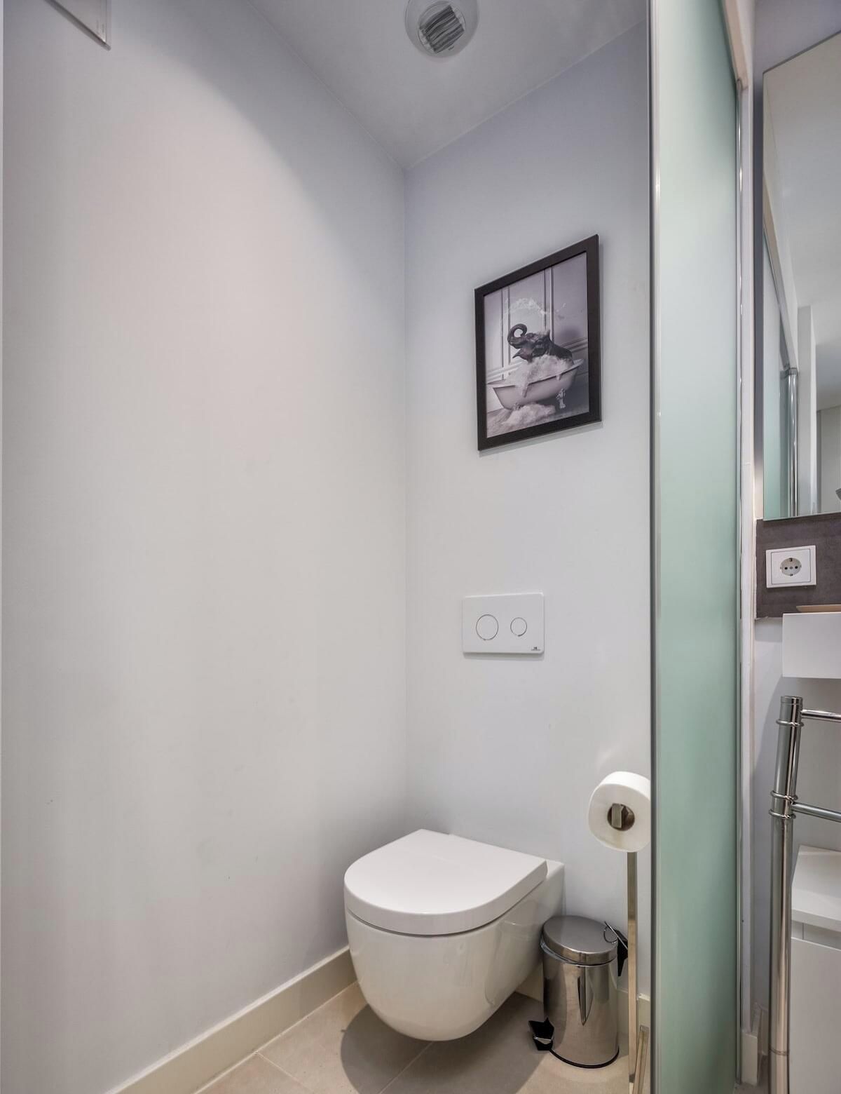 A small, modern bathroom with a toilet, a trash bin, a framed picture on the wall, and a light switch.