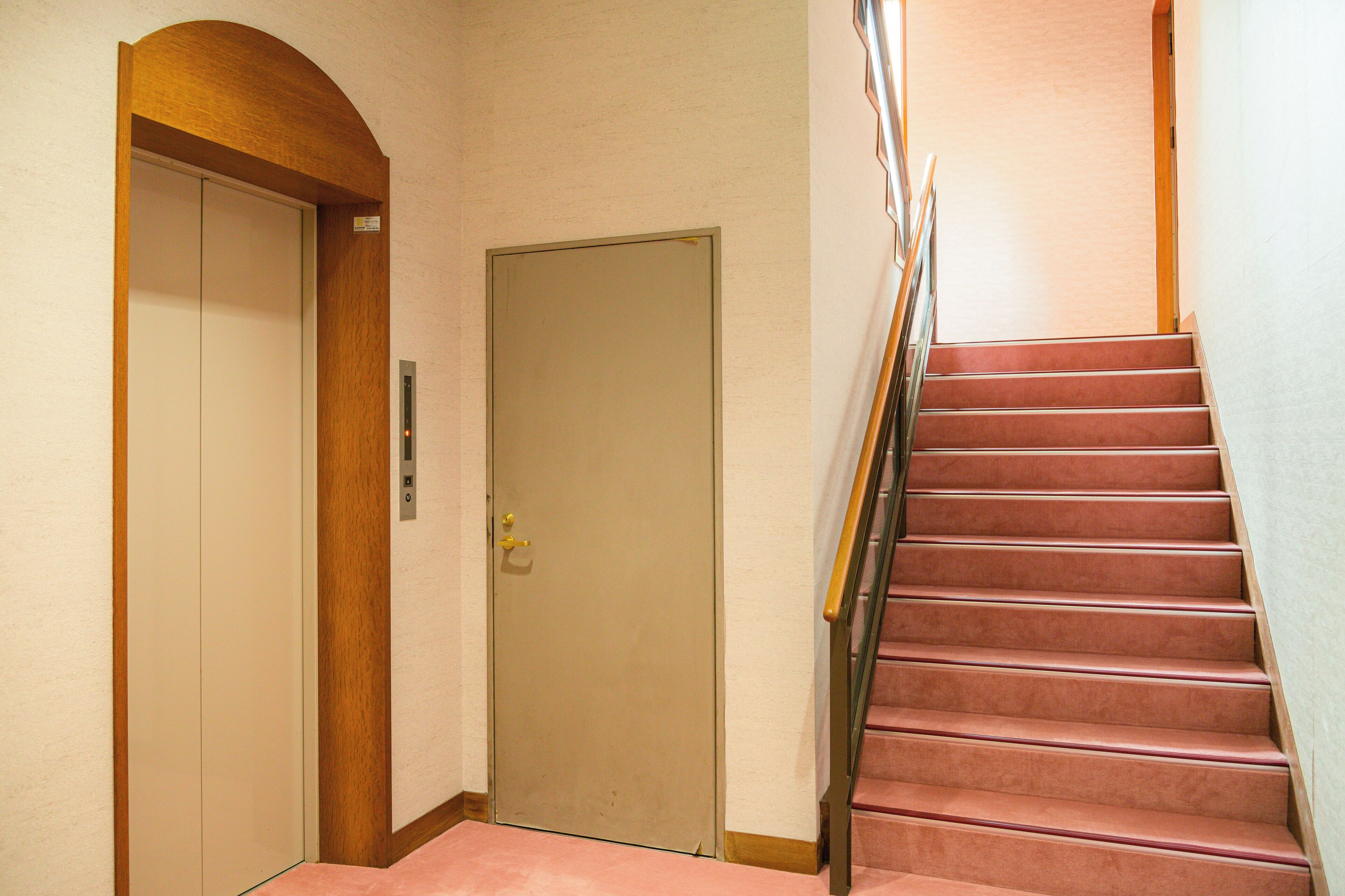 Un couloir avec une porte en bois, un ascenseur fermé et un escalier doté d’une rampe en métal.