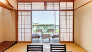 Una tradizionale stanza giapponese con pavimento in tatami, un tavolino con runner, due sedie e vista su una collina attraverso delle porte scorrevoli shoji.