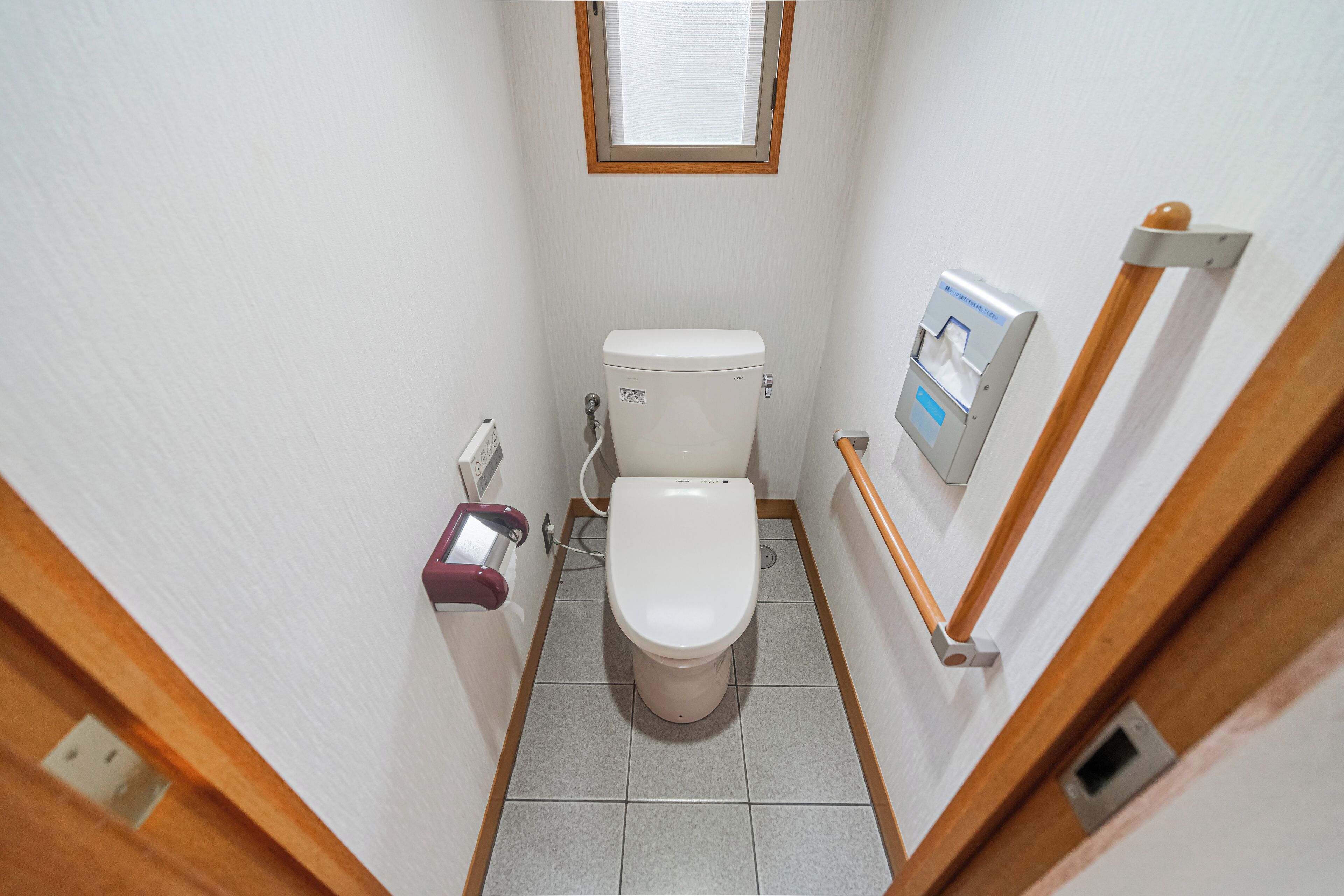A small, narrow bathroom with a toilet, a hand dryer, and a window above the toilet.