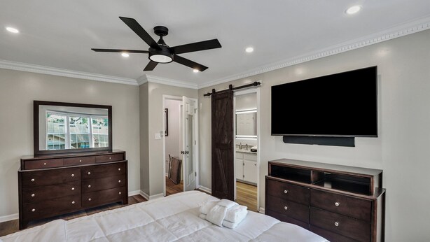 Un dormitorio con una cama, un aparador de madera oscura, un ventilador de techo, un televisor montado en la pared y una puerta que da a un baño.