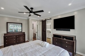 Un dormitorio con una cama, un aparador de madera oscura, un ventilador de techo, un televisor montado en la pared y una puerta que da a un baño.