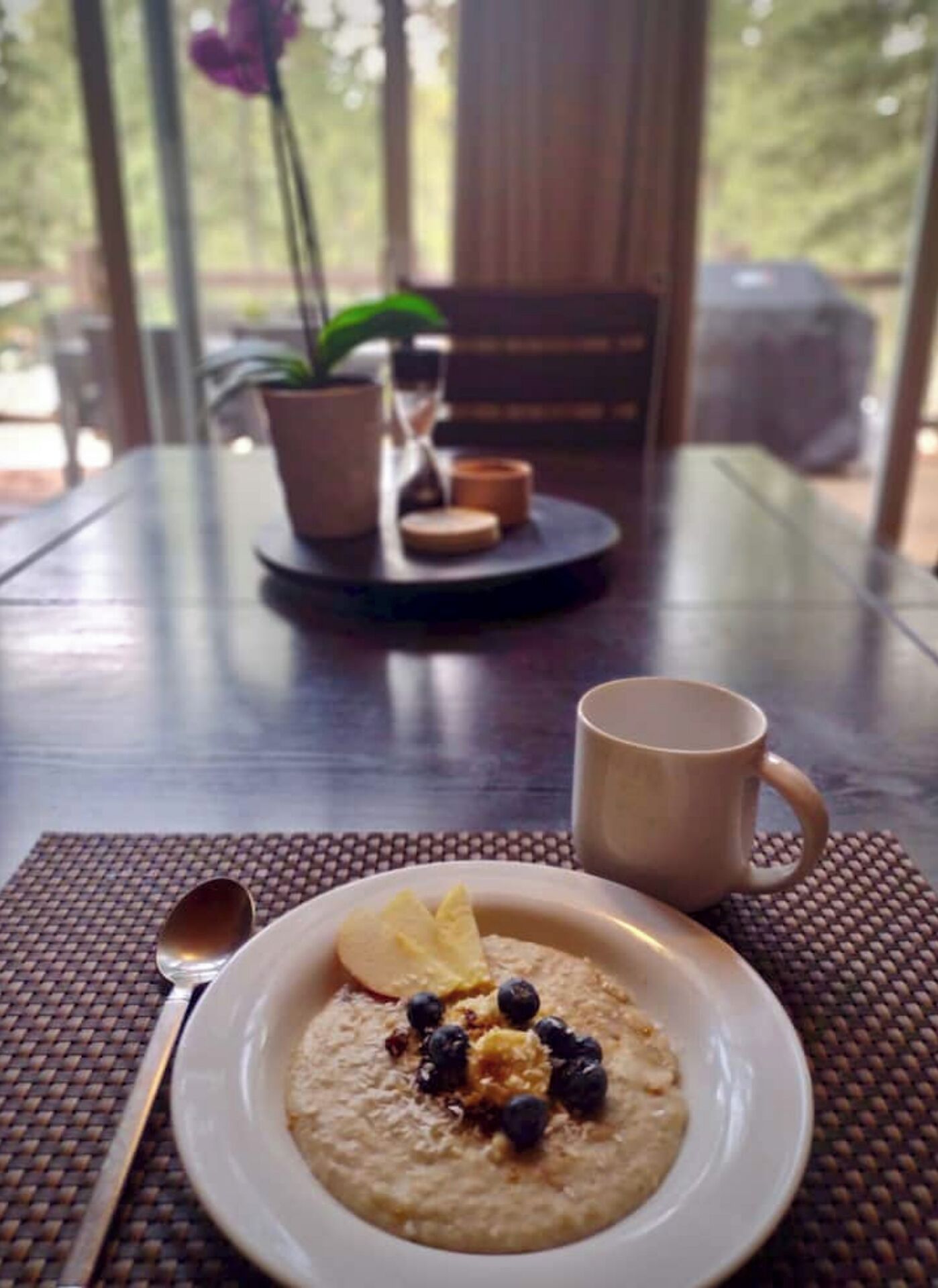 A dining table with a plate of oatmeal topped with blueberries and banana slices, a spoon, and a mug.