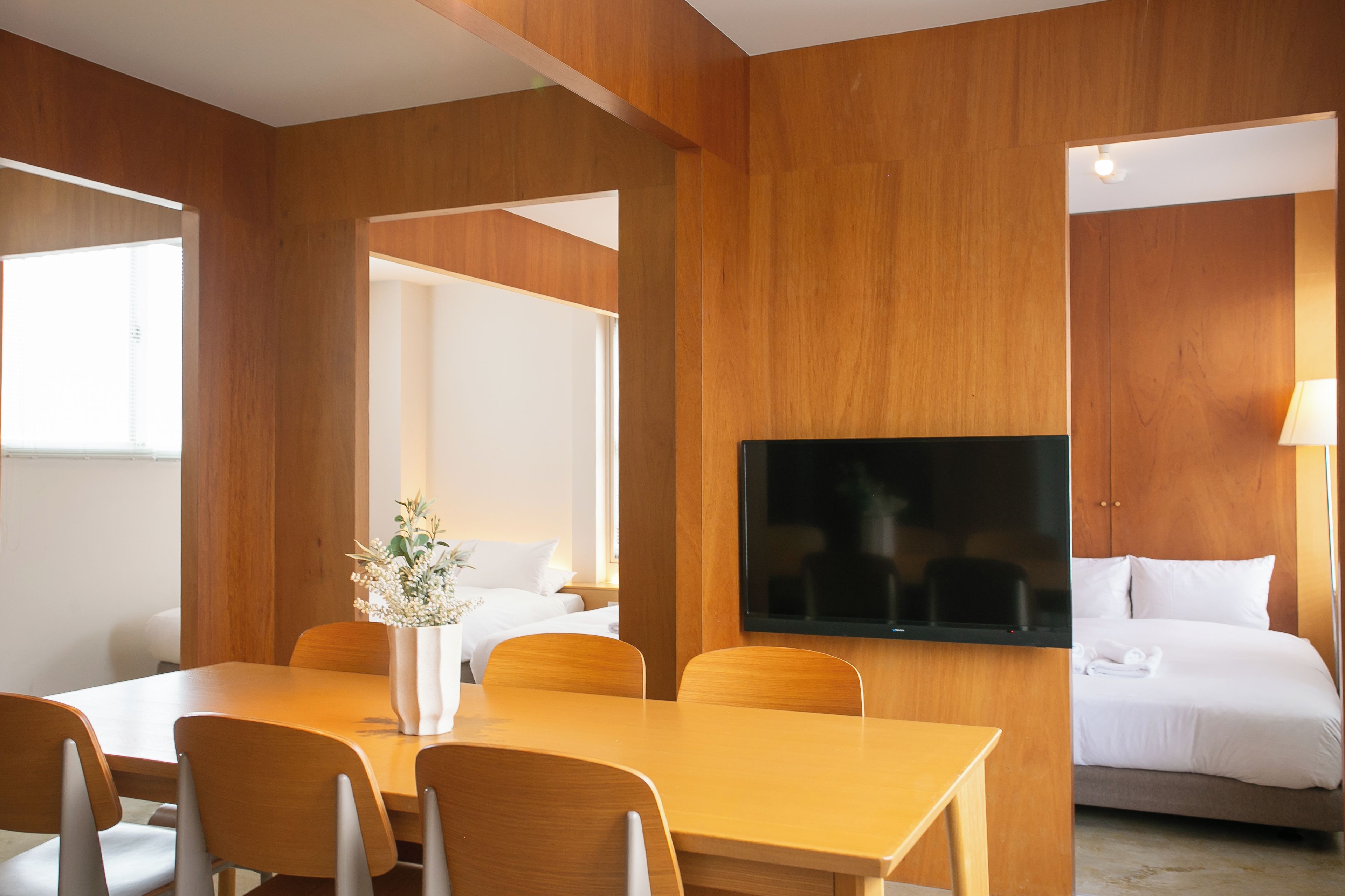 A hotel room with a wooden wall panel, a flat-screen TV, a bed, a dining table with chairs, and a vase with flowers.
