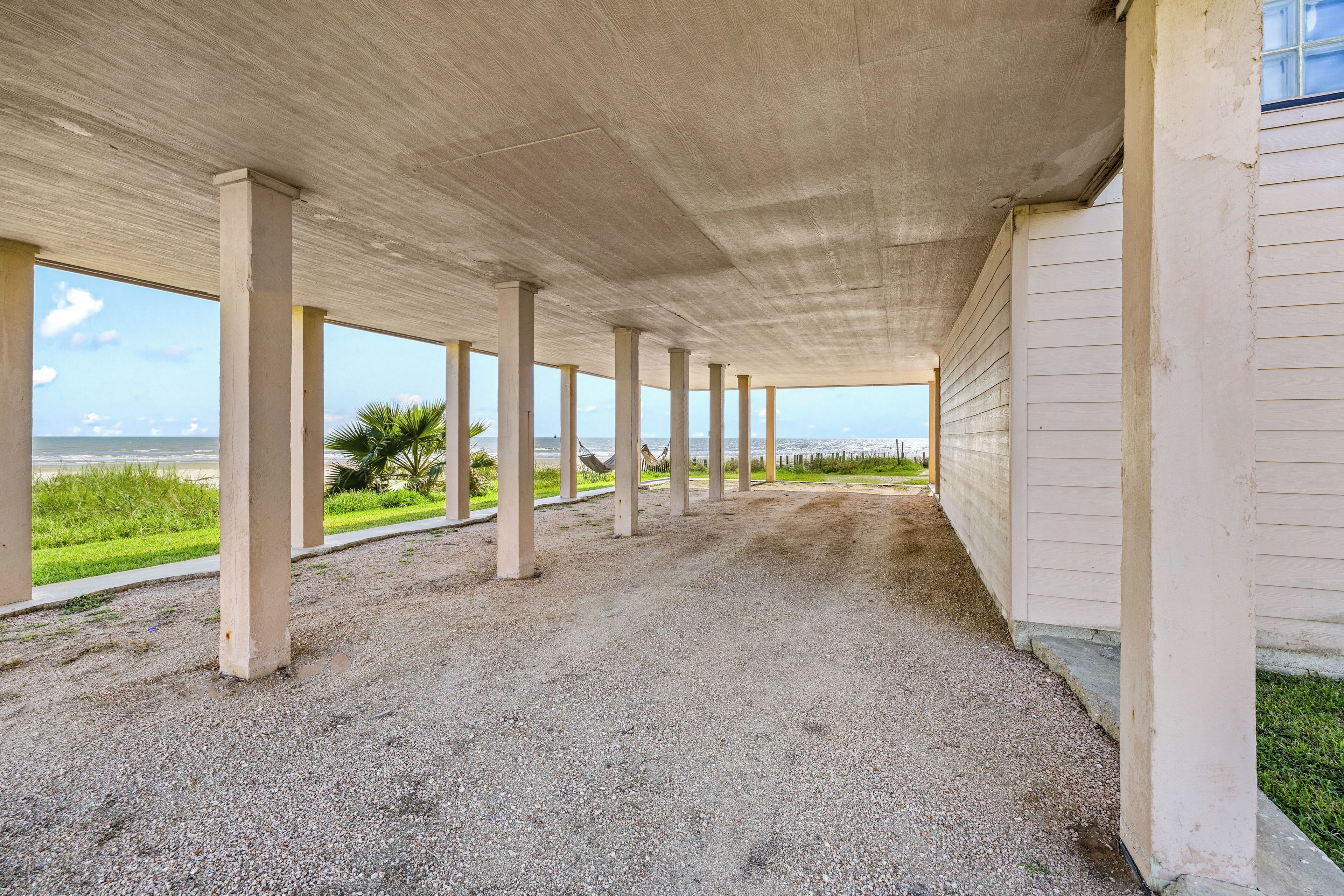Uma área externa coberta com pilares de concreto, piso de cascalho e vista para a praia e o oceano.