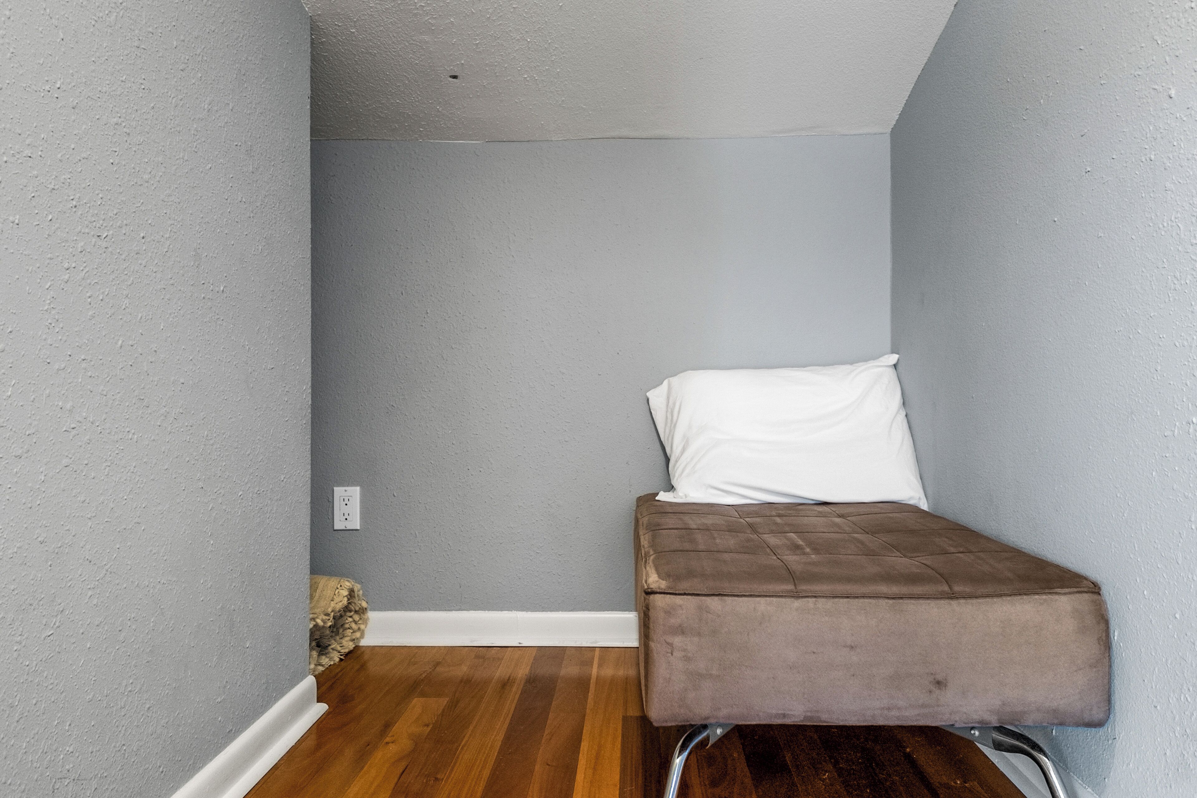 Una habitación pequeña y estrecha con suelo de madera, un banco marrón acolchado y un cojín blanco.