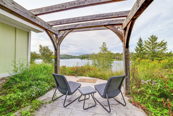 En overdækket terrasse med to fletstole og et lille bord, med udsigt over en sø og grønt område.