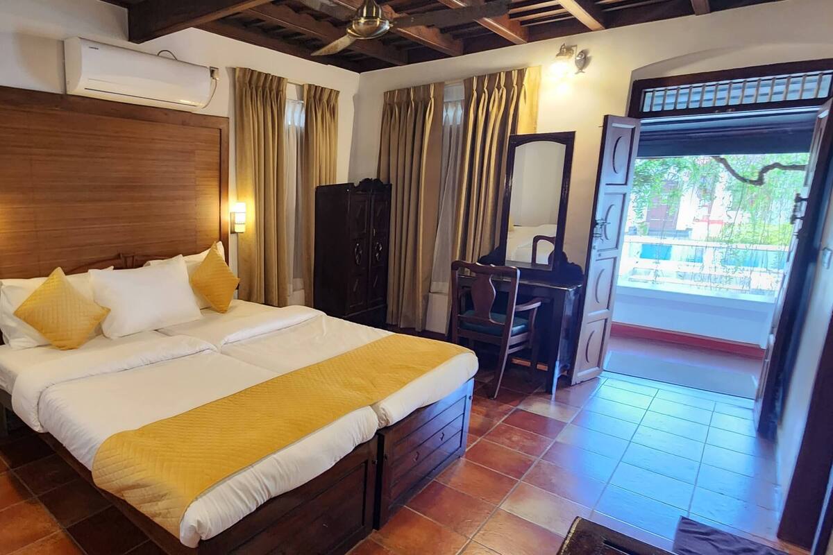Un dormitorio con cabecera de madera, cama con ropa de cama blanca y amarilla, mesita de noche, unidad de aire acondicionado, armario y vistas a un área exterior por una puerta.