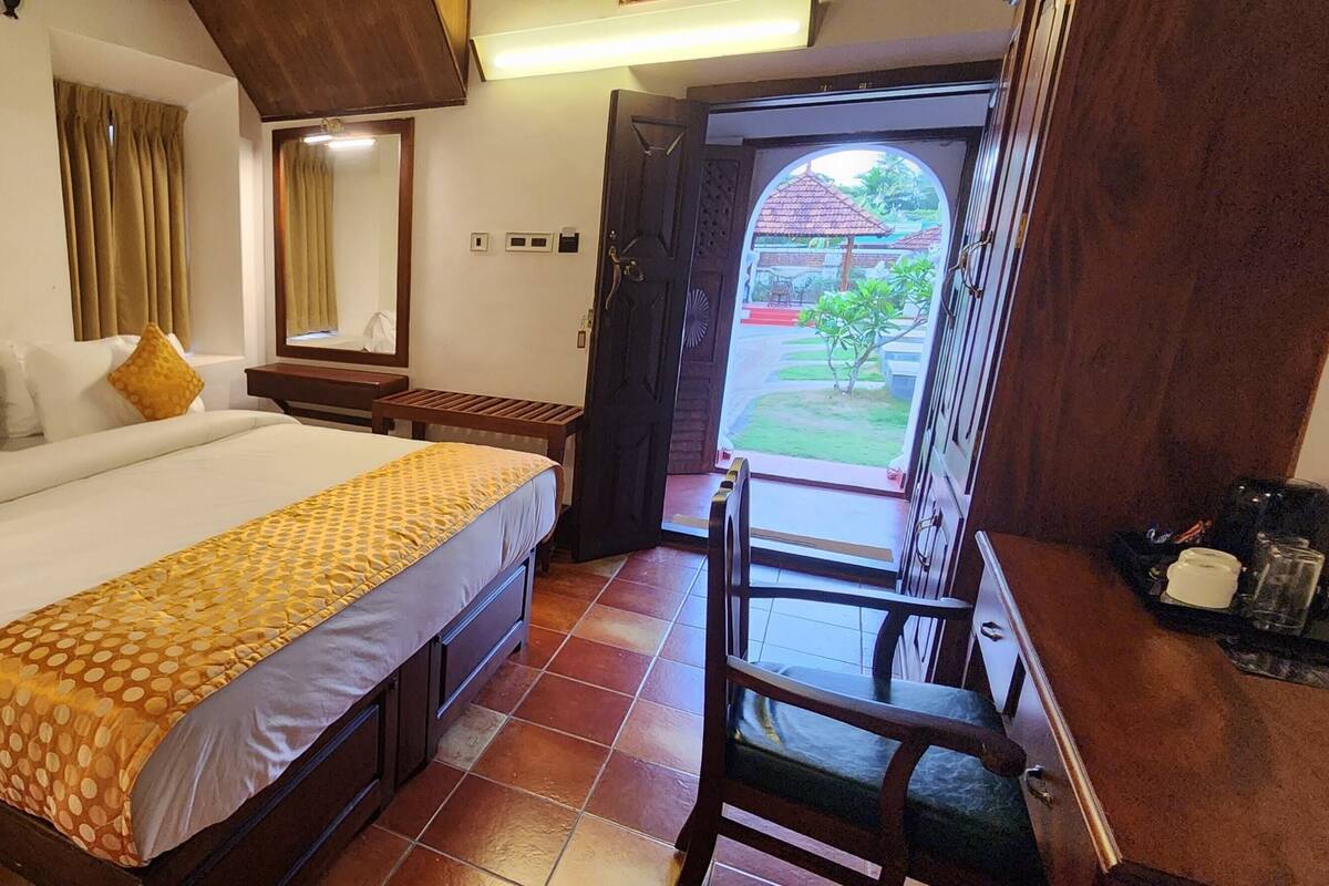 Habitación de hotel con una cama grande, un escritorio de madera, una silla, un espejo y vistas al jardín a través de una puerta abierta.