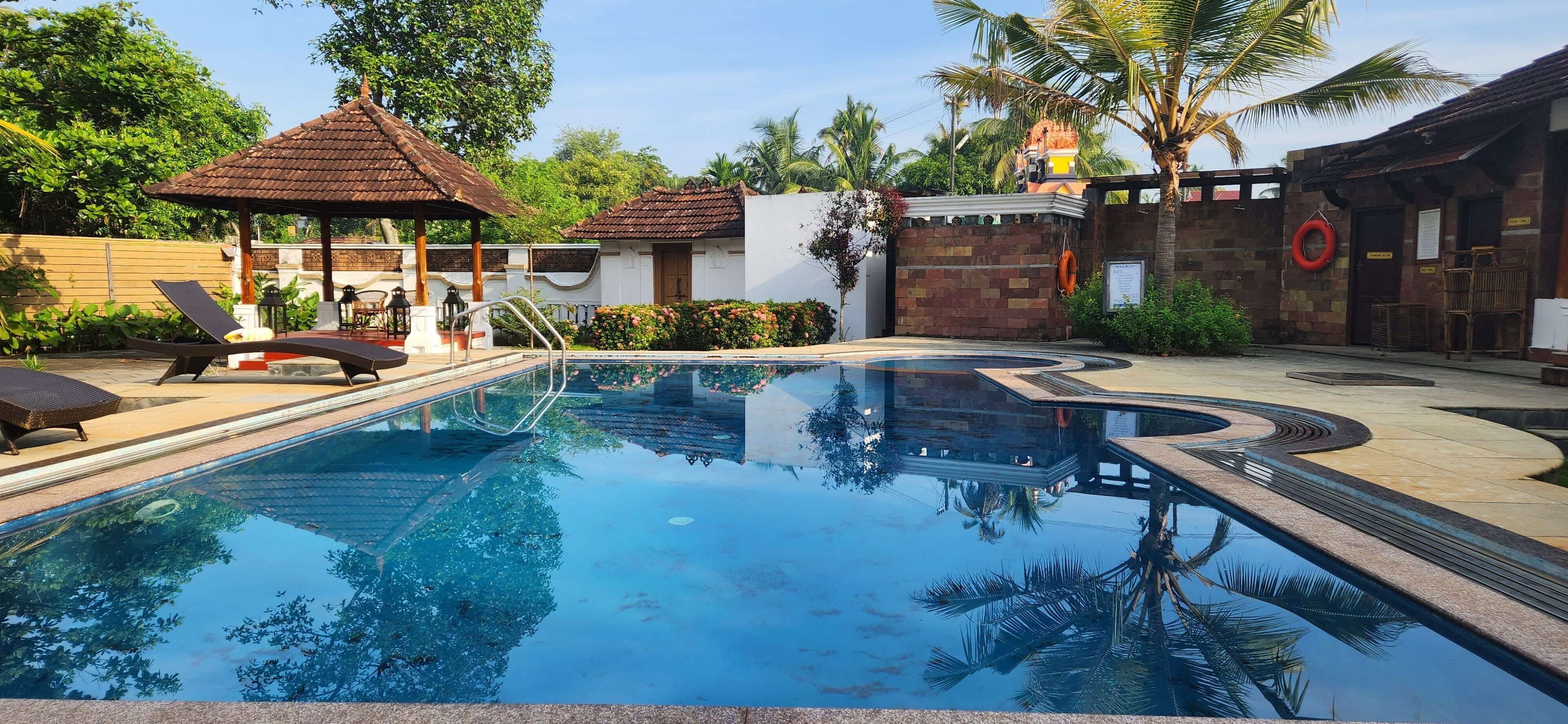 A swimming pool with lounge chairs, a gazebo, and a brick building with a palm tree in the background.