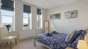 A bedroom with a bed, a chair, a bedside table, a lamp, and two windows with views of the outdoors. - Modern Apartment, Private Parking, Walk to Beach! (Westward Ho)