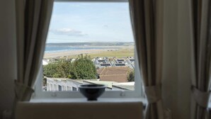 A view from a window with curtains, overlooking a coastal area with houses and a beach. - Modern Apartment, Private Parking, Walk to Beach! (Westward Ho)