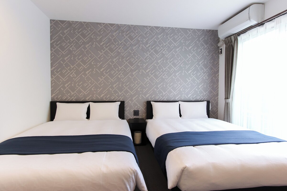 A hotel room with two beds, a grey patterned wall, a window with curtains, and a wall-mounted air conditioner.
